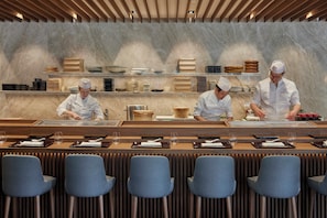 A restaurant with a marble wall, chefs preparing food, and a long dining table with chairs.