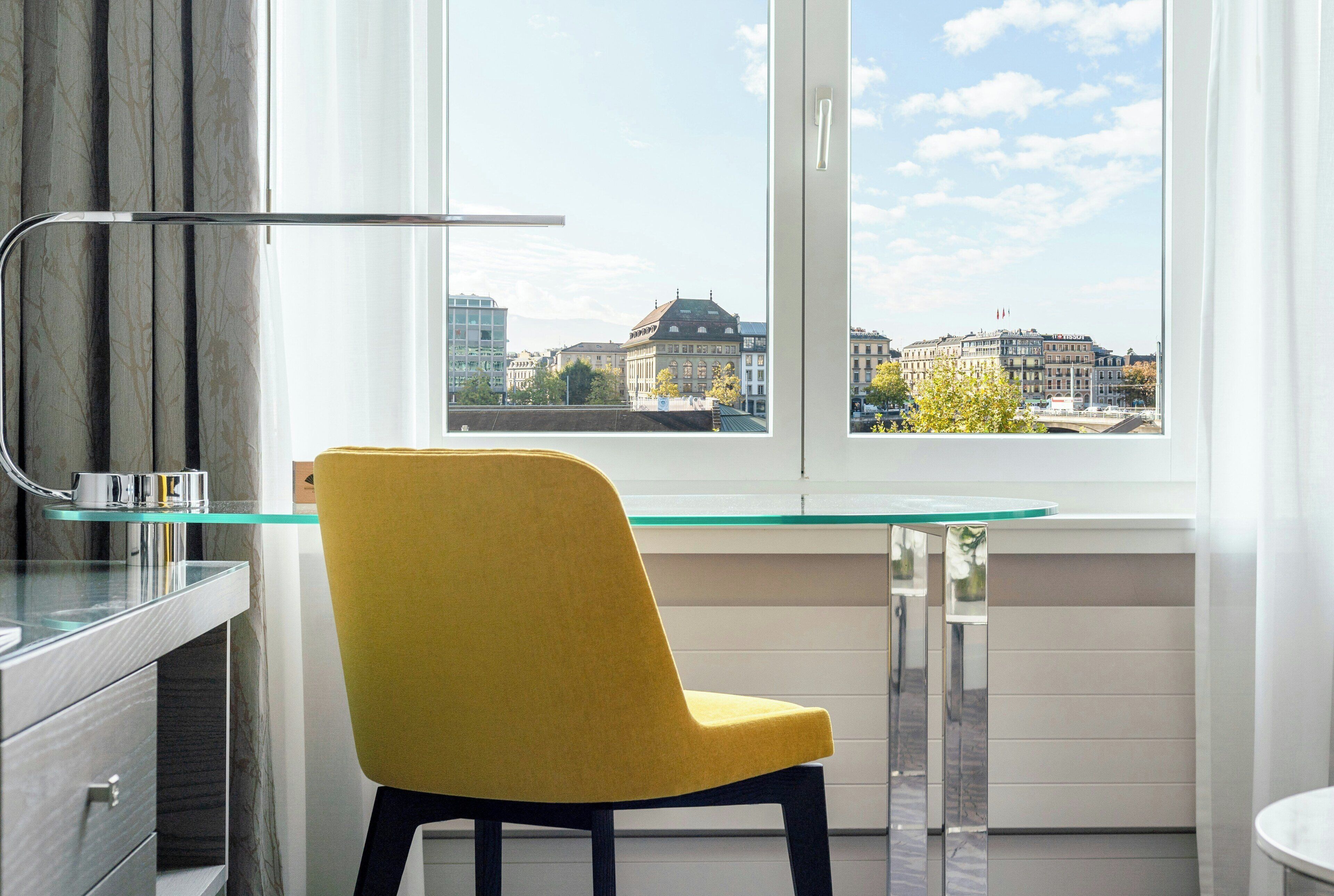Eine moderne Küche mit einem gelben Stuhl, einem Glastisch und Blick auf Stadtgebäude durch ein großes Fenster.