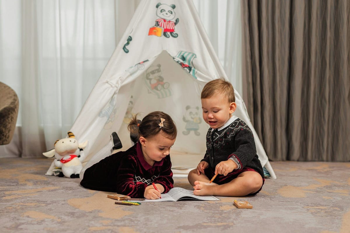 Two children sitting on a carpeted floor, one writing in a notebook and the other drawing, with a white tent featuring cartoon characters in the background.