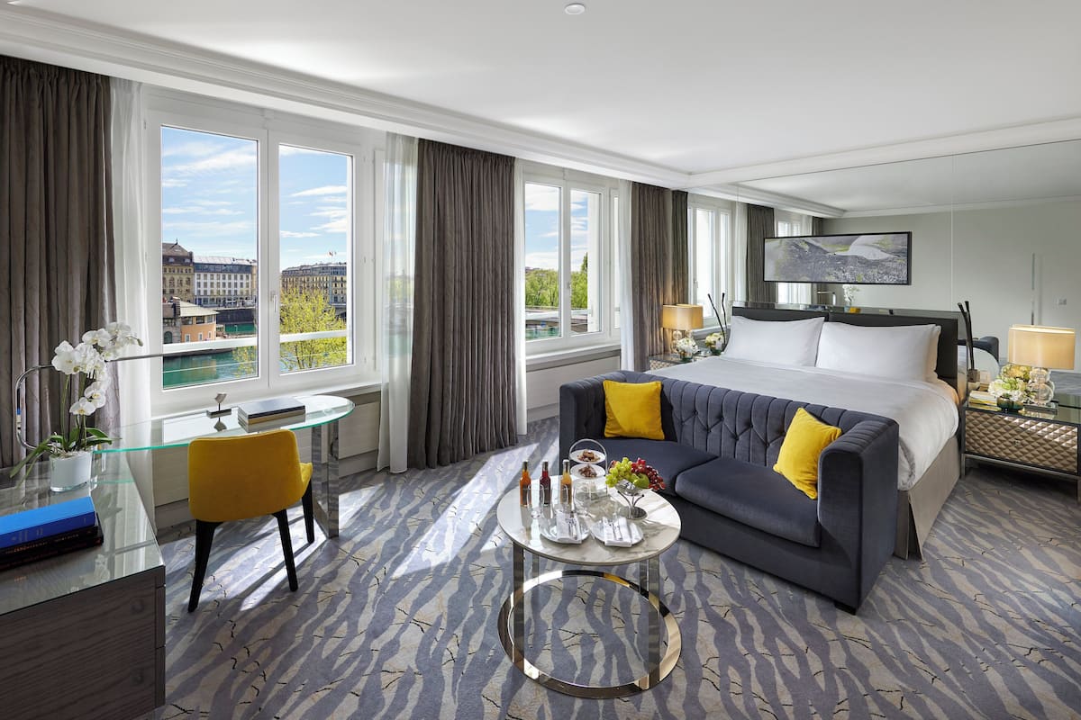 Habitación de hotel con cama, sofá, escritorio y vistas a edificios a través de amplias ventanas.