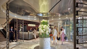 Un vestíbulo moderno con suelo de mármol, un arreglo floral central, una recepción y una zona con plantas, todo ello envuelto en cristal.