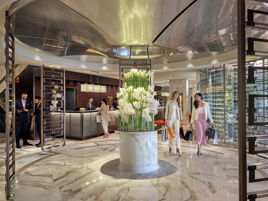 A modern lobby with marble flooring, a central floral arrangement, a reception desk, and a glass-enclosed area with plants.