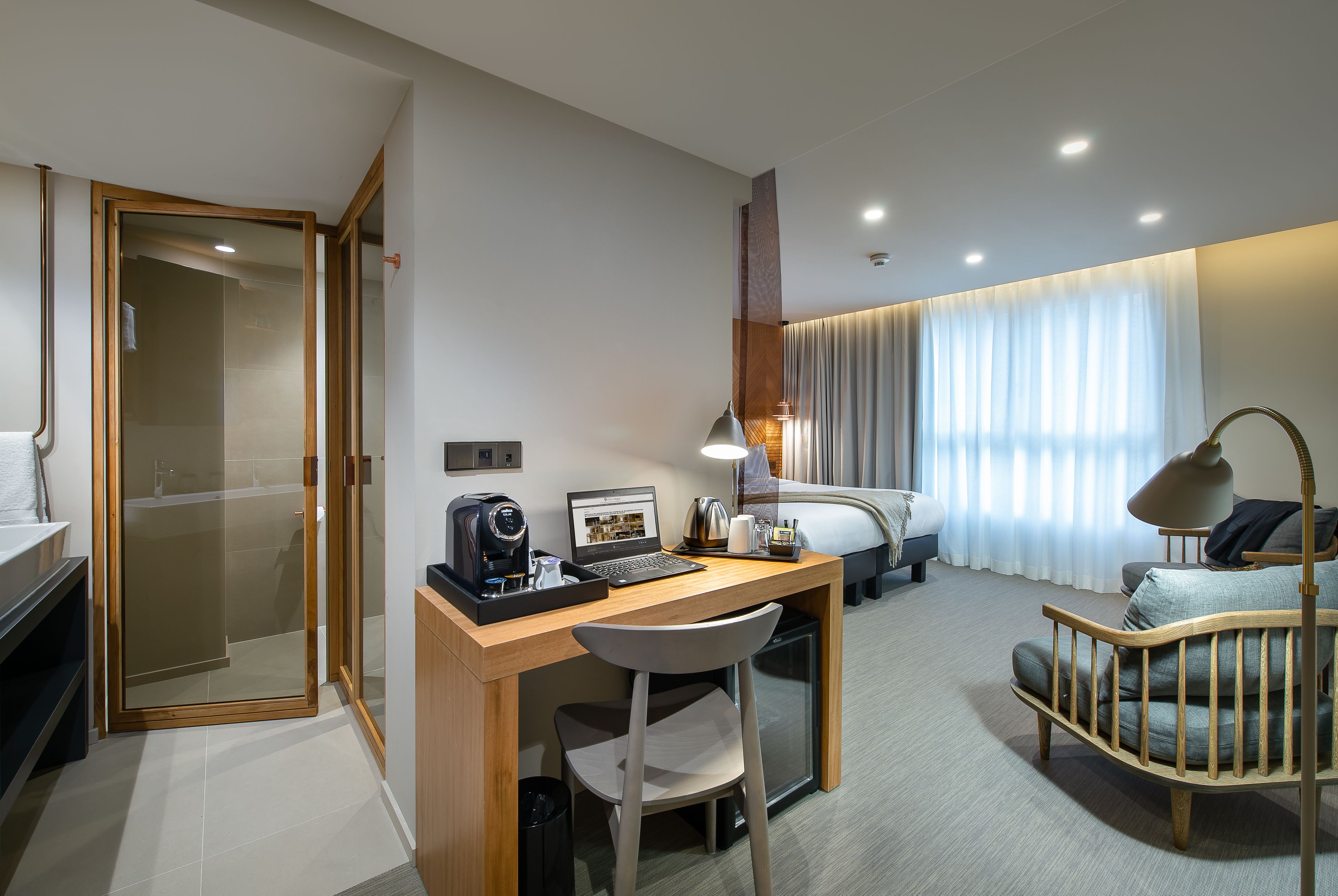 Ein Hotelzimmer mit Schreibtisch aus Holz, einem Laptop, einer Kaffeemaschine, einem Sessel, einem Babybett, einem Bett und einem Duschbereich.