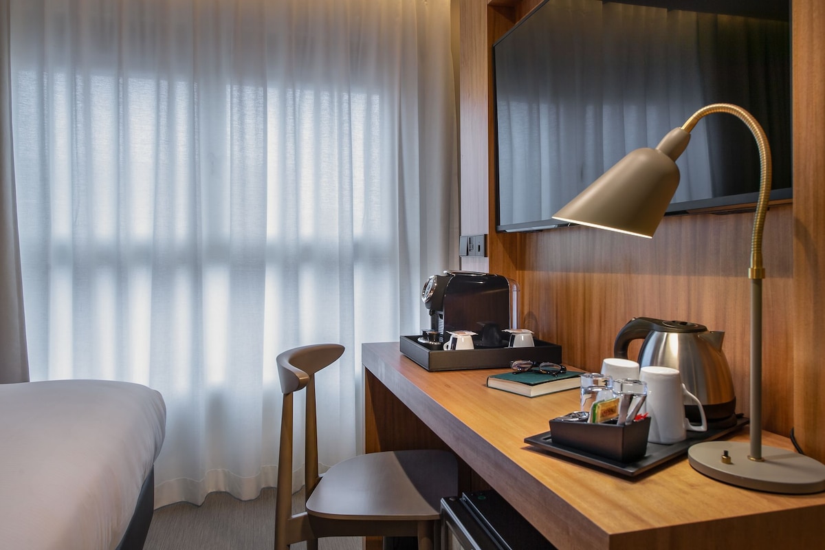 A hotel room with a wooden desk, a coffee maker, a kettle, a lamp, and a chair.