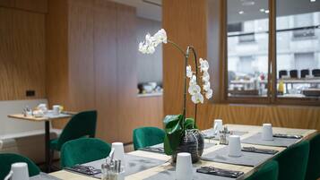 Un espace de restauration moderne avec des chaises vertes, une longue table dressée avec des tasses blanches, des couverts et une orchidée en pot.