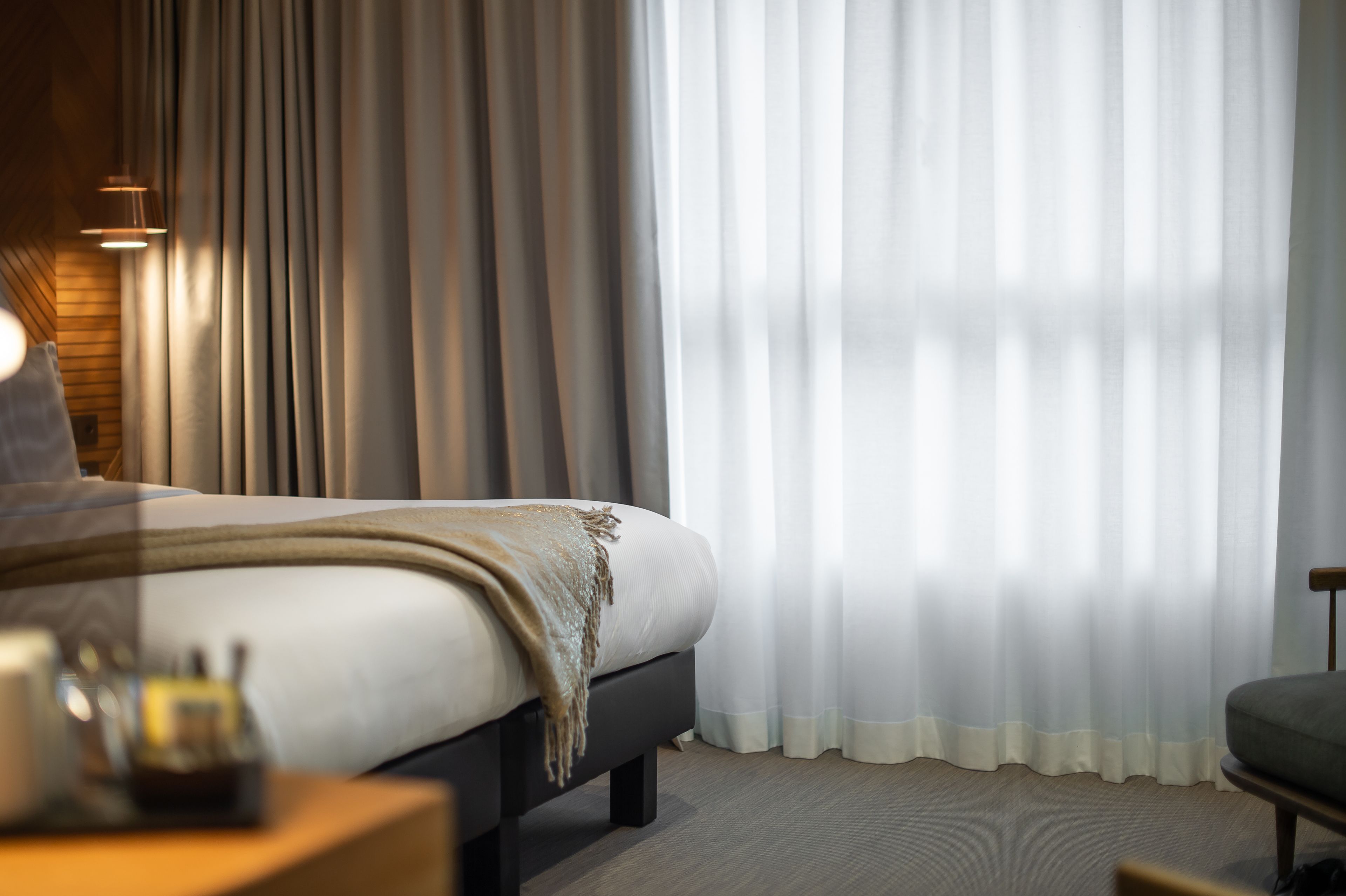 Una habitación de hotel moderna con una cama grande, una mesita de noche con lámpara y cortinas translúcidas.