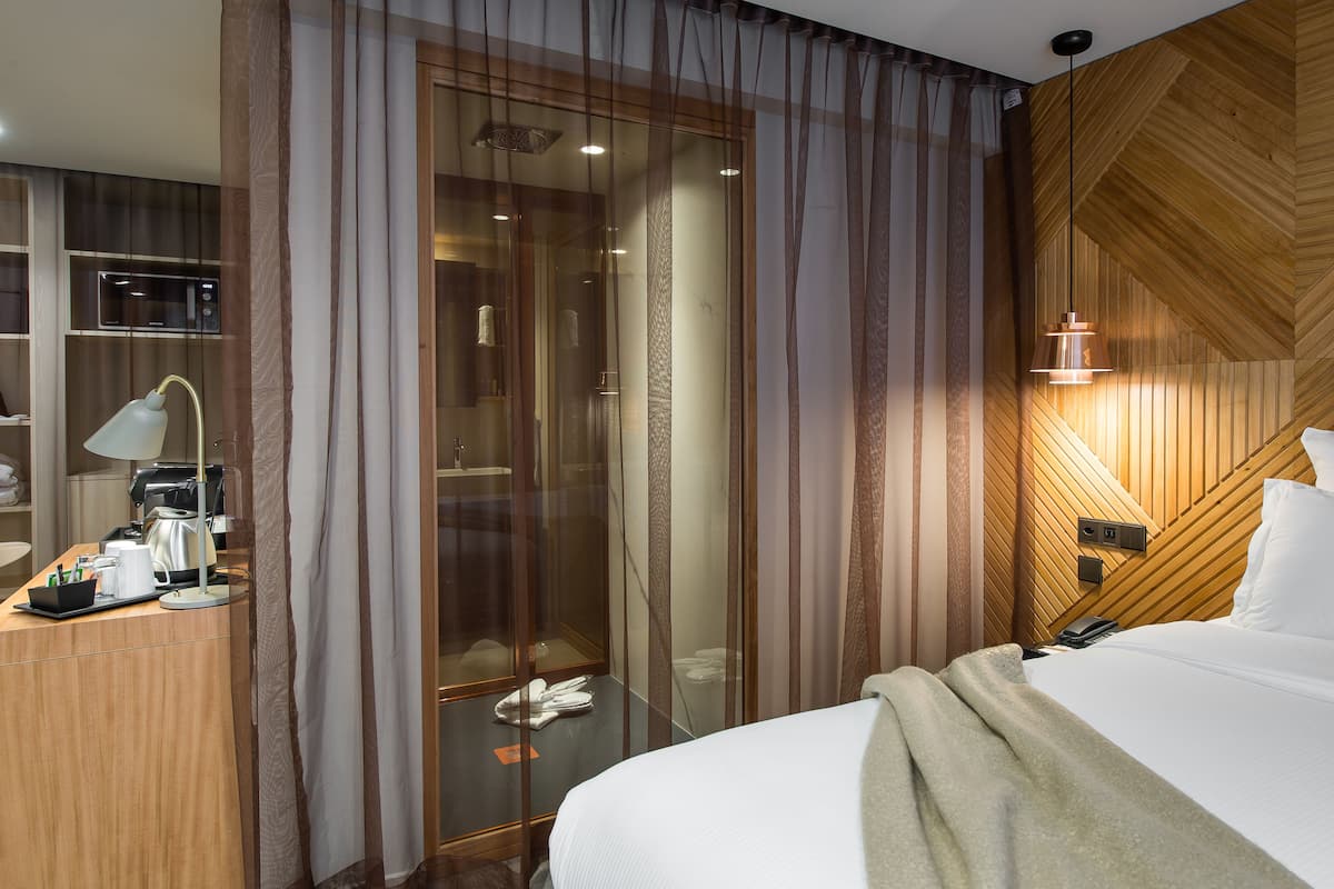 Hotellihuone, jossa on sänky, kirjoituspöytä lampun kanssa, kahvinkeitin ja suihkutila.
