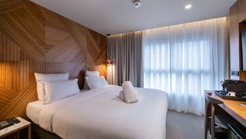A modern hotel room with a wooden accent wall, a large bed with white linens, a desk with a coffee maker, and a view of the outdoors through sheer curtains.