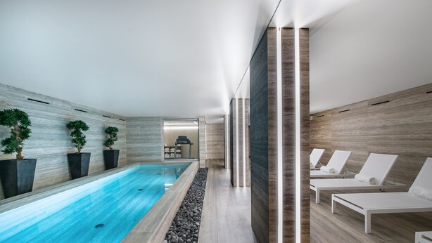 Un área de piscina interior moderna con una piscina larga y estrecha, sillas de descanso y plantas en macetas a lo largo de las paredes.