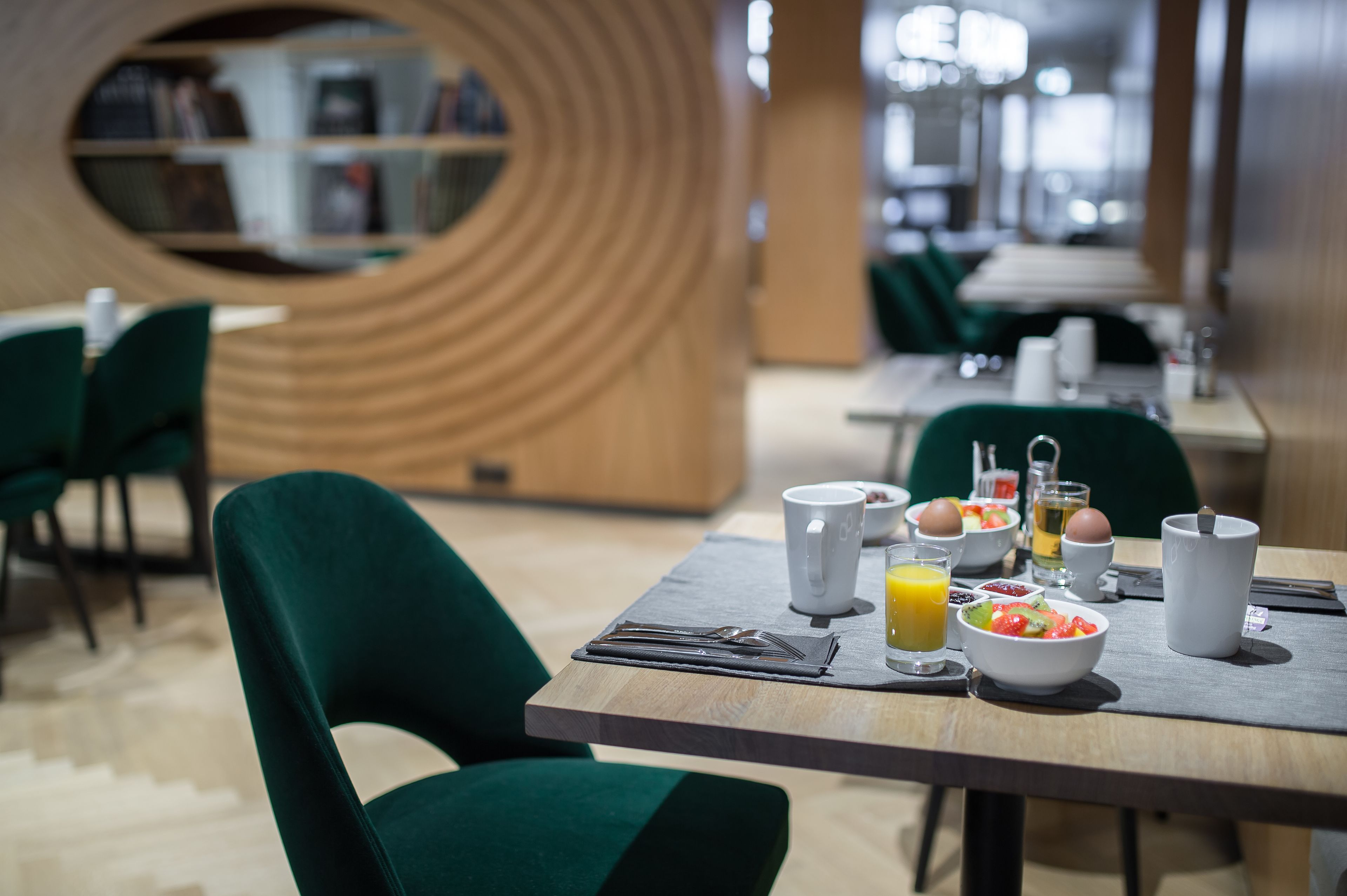 Un espace repas moderne avec un mur incurvé en bois, des chaises recouvertes d’un tissu vert et une table de petit-déjeuner agrémentée d’un saladier de fruits, d’œufs et d’un verre de jus d’orange.