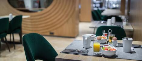 Un espace repas moderne avec un mur incurvé en bois, des chaises recouvertes d’un tissu vert et une table de petit-déjeuner agrémentée d’un saladier de fruits, d’œufs et d’un verre de jus d’orange.