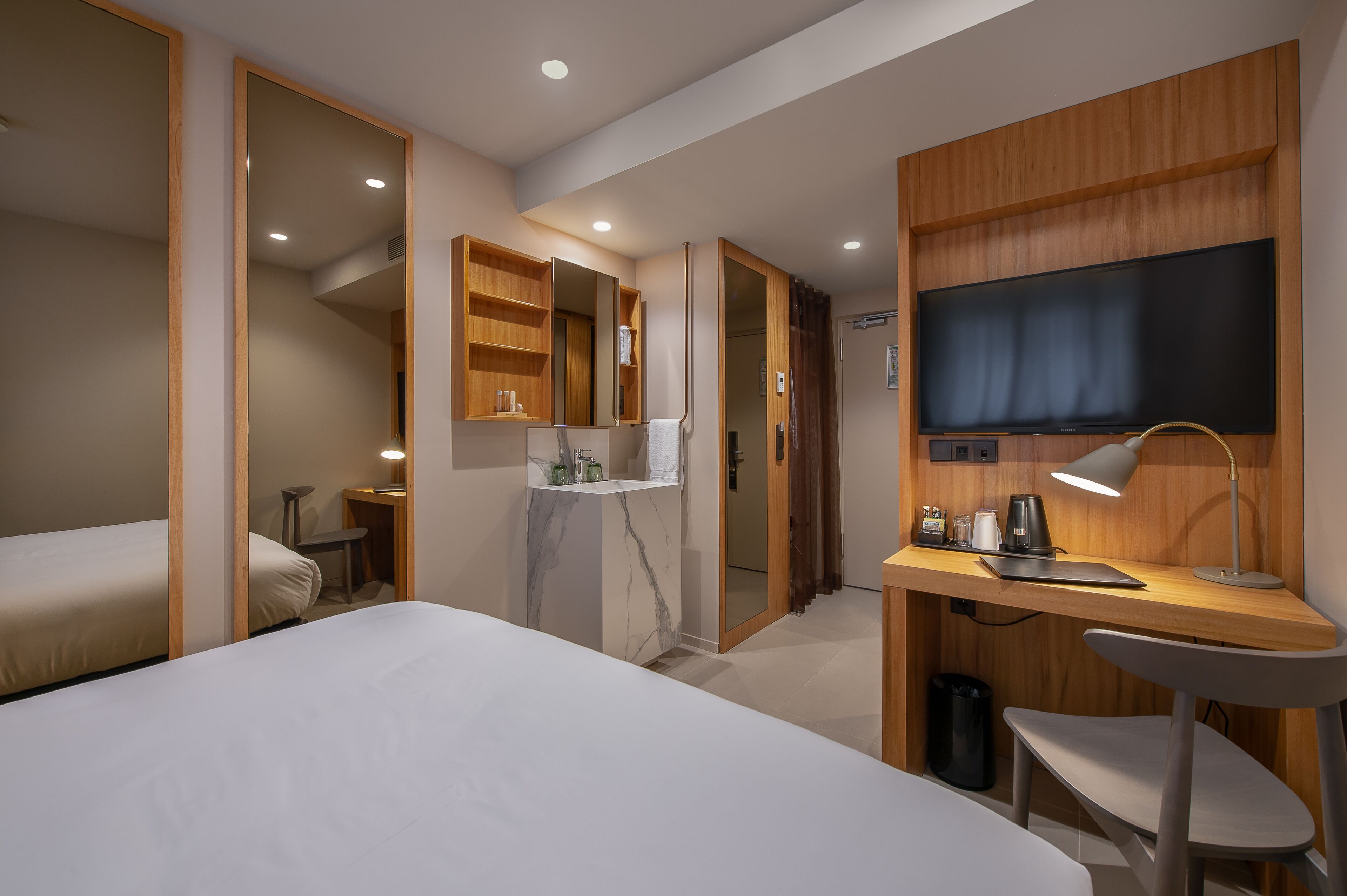 Una habitación de hotel moderna con cama, escritorio con silla, televisión y baño con espejo y estantes.