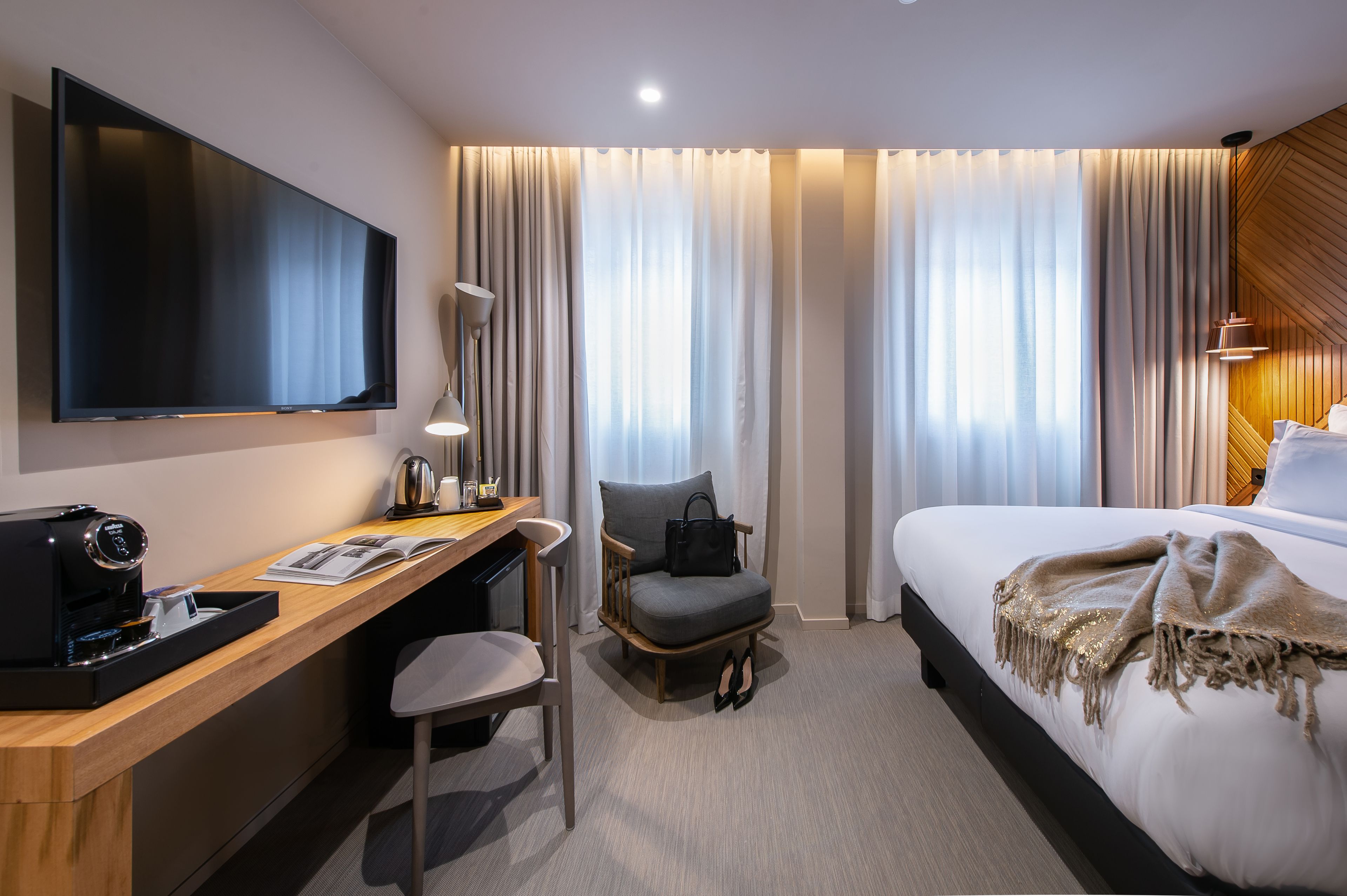 Ein Hotelzimmer mit Flachbildfernseher, Schreibtisch mit Kaffeemaschine, Stuhl, Bett mit Decke und großen Fenstern mit Vorhängen.