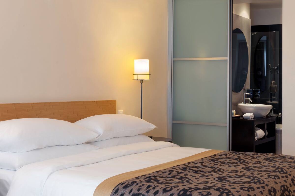 A neatly made bed with white linens and a patterned throw blanket, a bedside lamp, and a modern bathroom with a large mirror and sink.