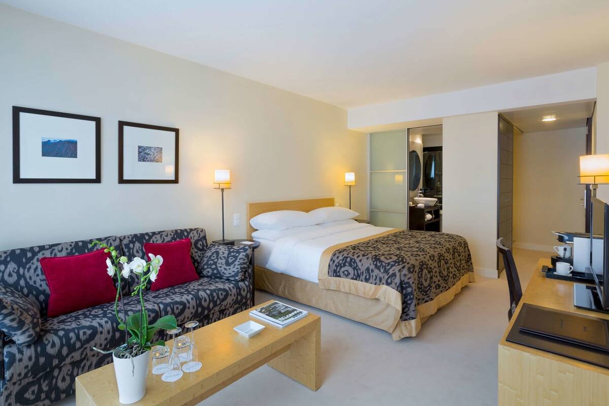 A hotel room with a bed, a sofa, a coffee table, a TV, and a bathroom visible through an open door.