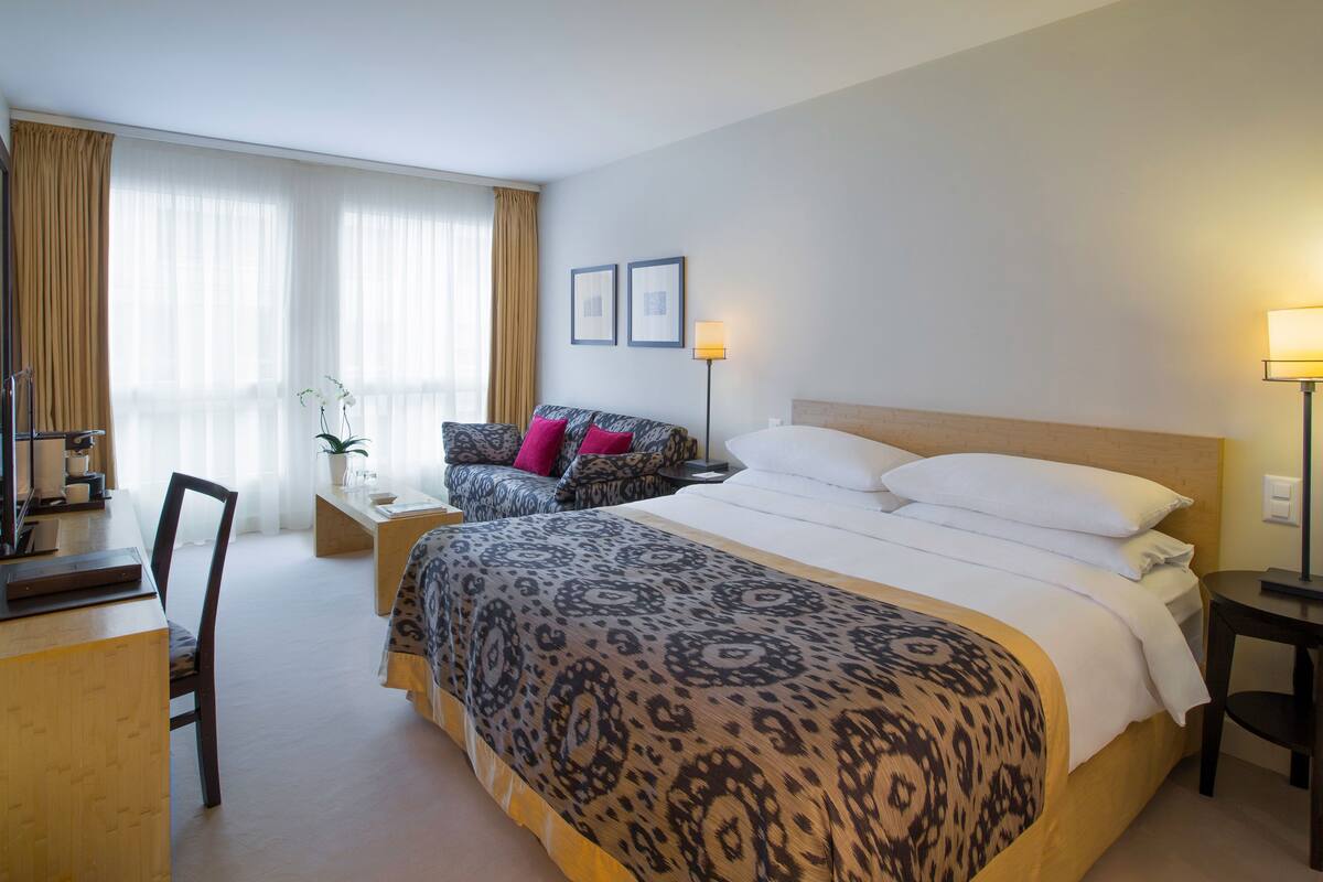 A hotel room with a large bed, a desk, a chair, a sofa, and two bedside tables.