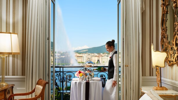 A room with a balcony, a table with a tea set, and a view of a city and mountains. - Hotel d'Angleterre Geneva (Geneva)