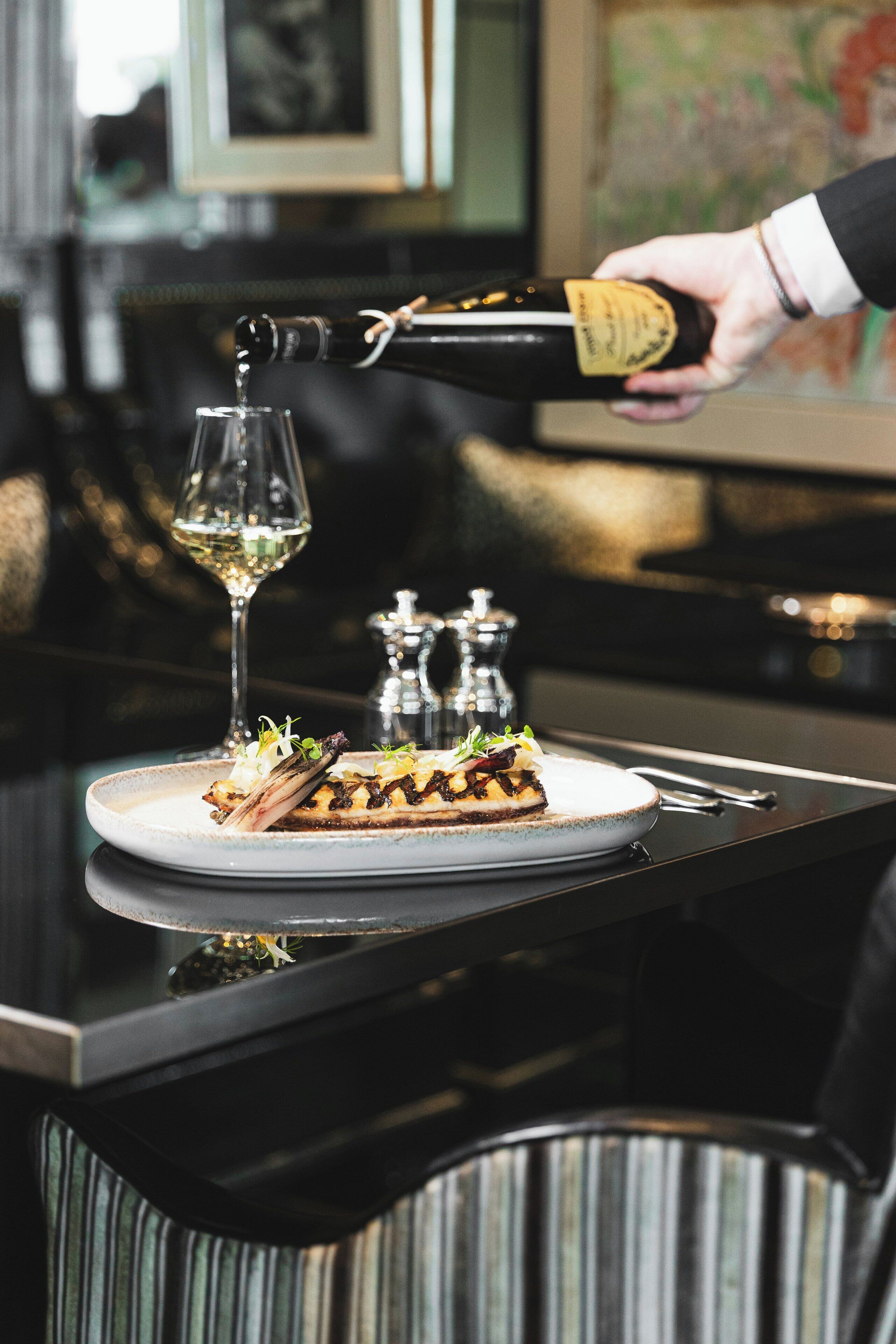 Una persona sirve vino en una copa, un plato de comida y saleros y pimenteros sobre una mesa.