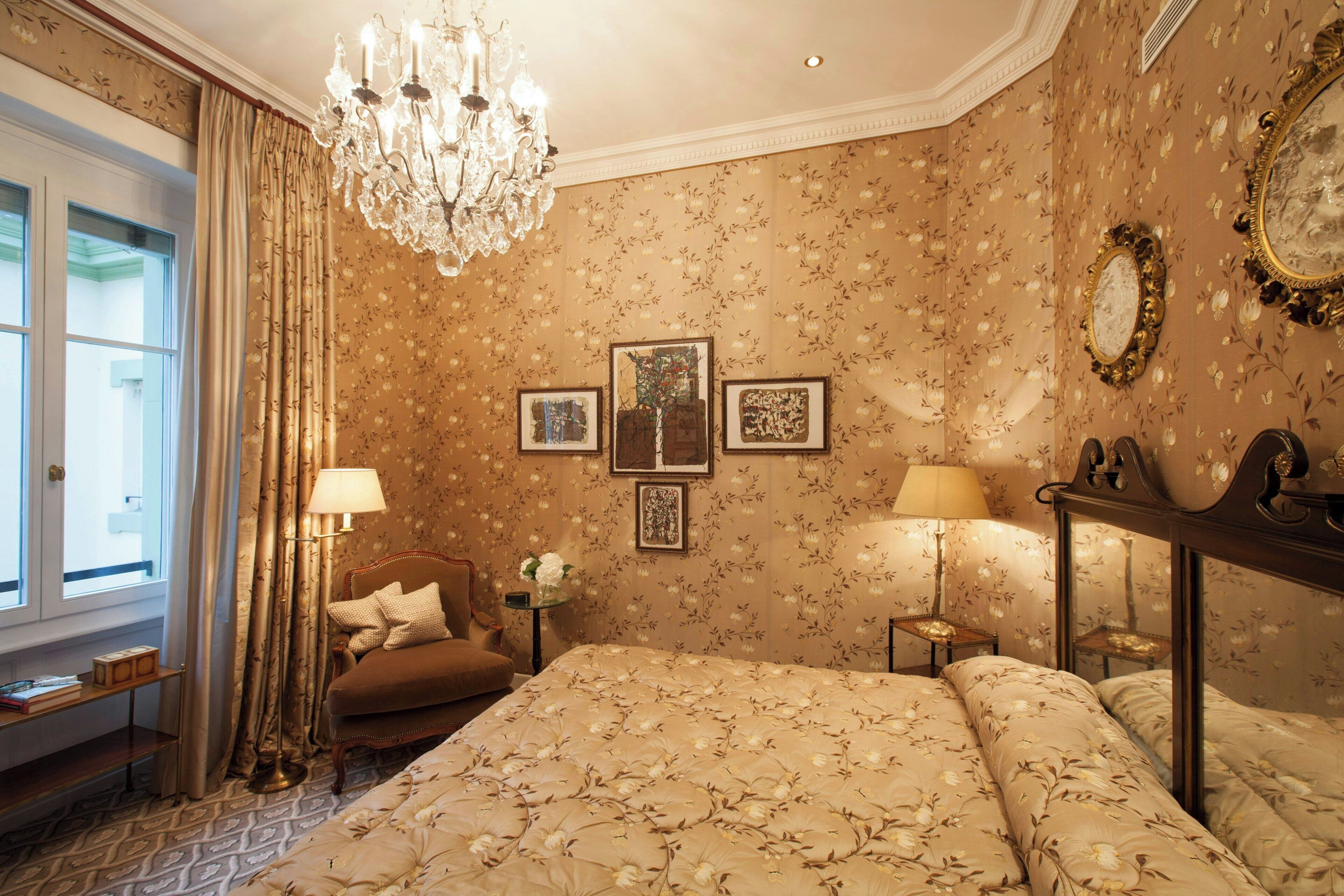Ein Hotelzimmer mit einem Bett, einer Chandelier, einem Fenster mit Vorhängen und gerahmten Bildern an der Wand.