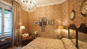 A hotel room with a bed, a chandelier, a window with curtains, and framed pictures on the wall.
