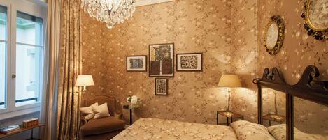 Habitación de hotel con cama, candelabro, ventana con cortinas e imágenes enmarcadas en la pared.