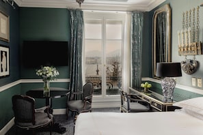 A hotel room with a bed, two chairs, a table with a vase, a lamp, a TV, and a large window with curtains. - Hotel d'Angleterre Geneva (Geneva)