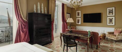 A hotel room with a large window, a chandelier, a television, a sofa, a desk, and a dresser.