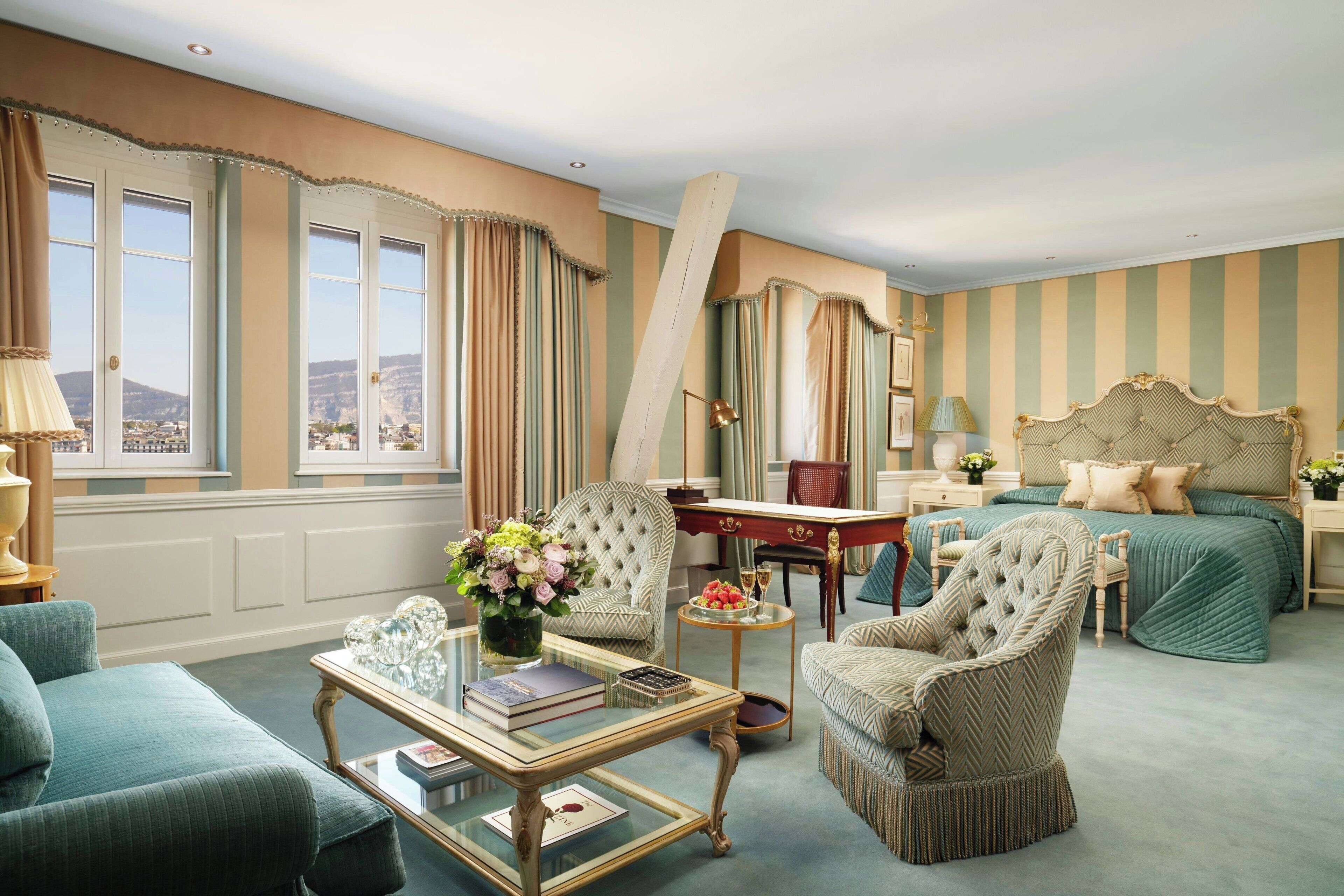A hotel room with a large bed, two armchairs, a coffee table, a desk, and a view of the city through large windows.