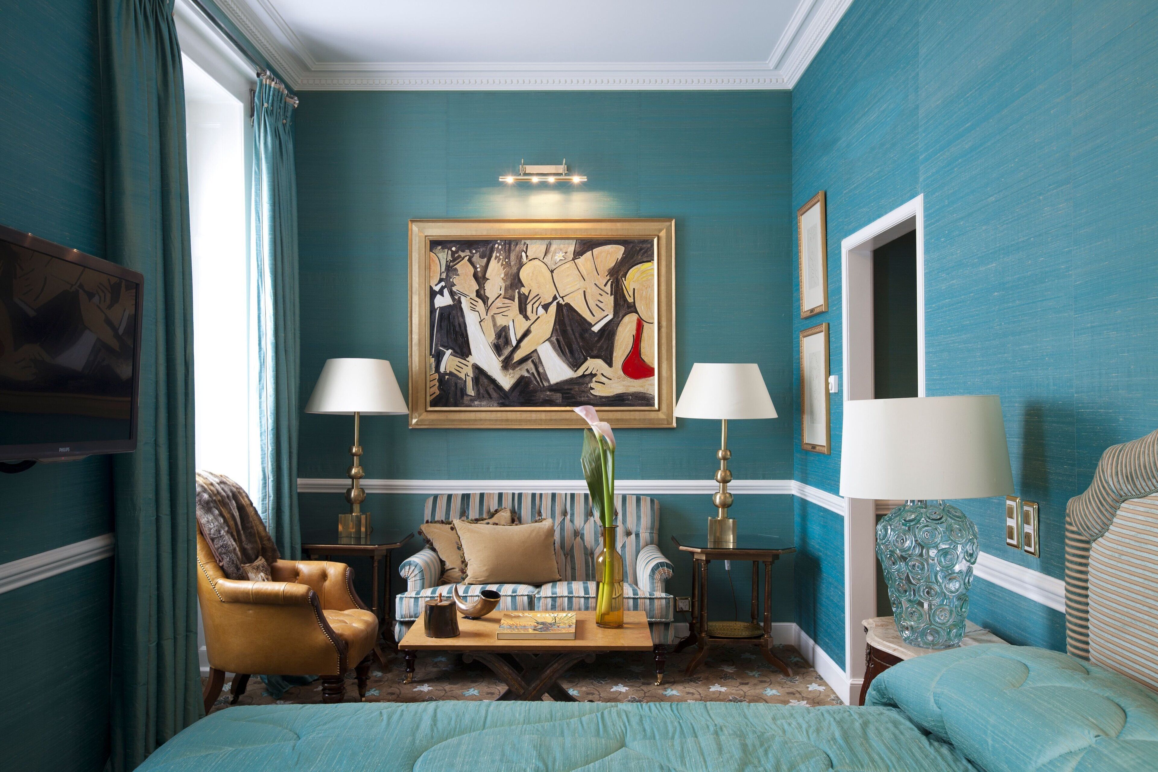 A room with teal walls, a large abstract painting, a sofa, two armchairs, a coffee table, and a bed.