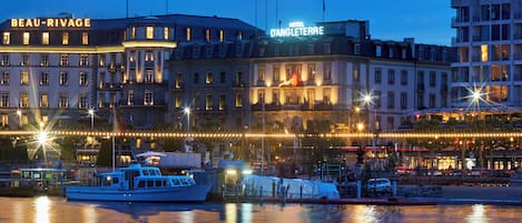 A waterfront hotel with illuminated signage, a boat docked nearby, and a reflection on the water.