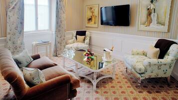 Una sala de estar con un sofá marrón, una mesa de centro blanca y dos sillones con estampado. Un televisor de pantalla plana está montado en la pared, y hay cortinas con motivos florales.