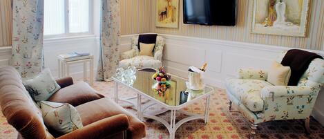 Ein Wohnzimmer mit einem braunen Sofa, einem weißen Couchtisch und zwei gemusterten Sesseln. Ein Flachbildfernseher ist an der Wand montiert, und es gibt Vorhänge mit Blumenmuster.