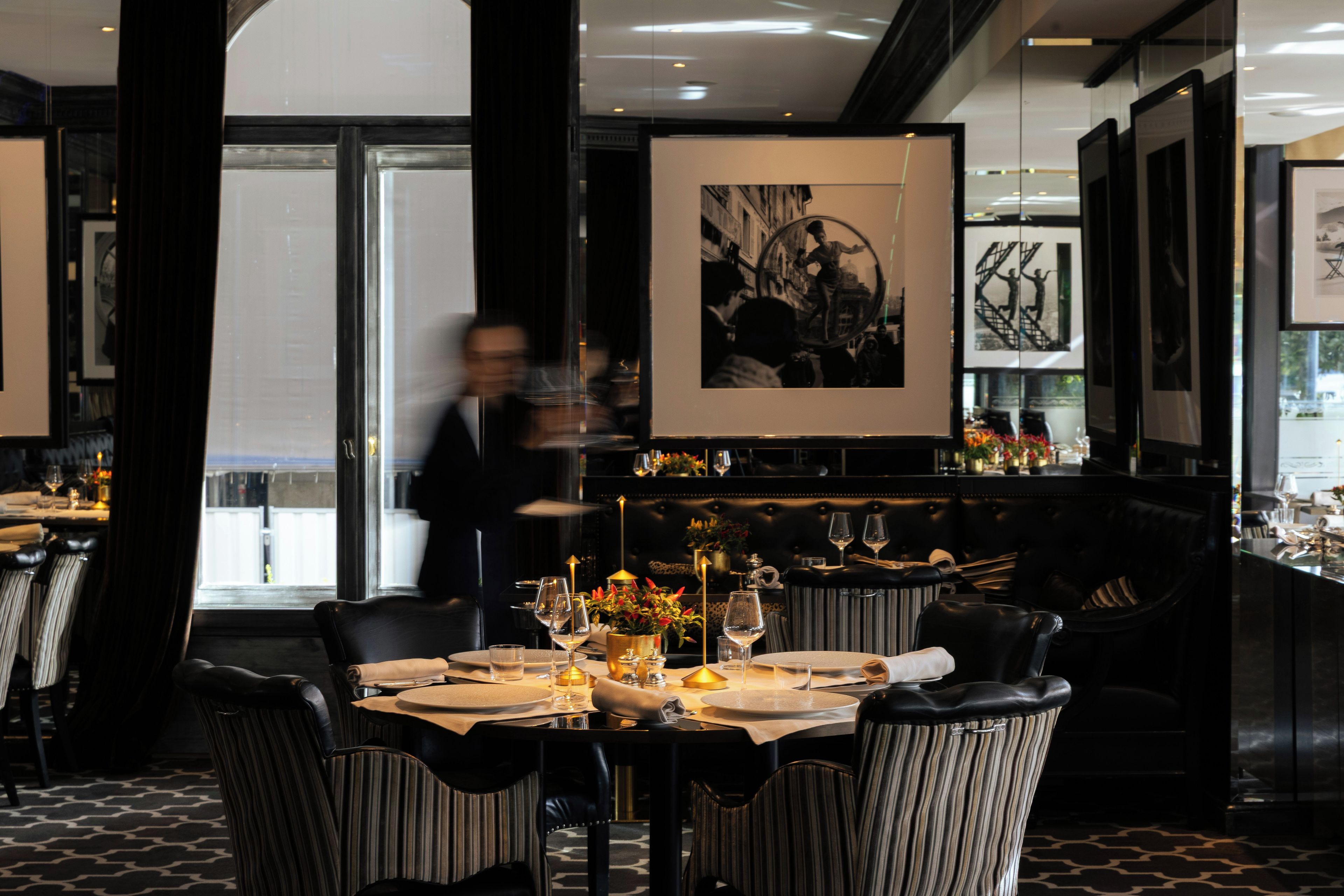 Interior de restaurante com mesa posta para dois, cercada por cadeiras estofadas.