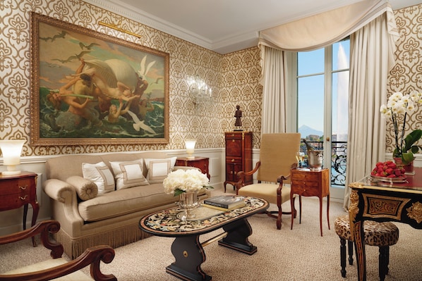 Una habitación lujosa con un cuadro grande y ornamentado, un sofá, una mesa de centro con un jarrón y vistas de la ciudad a través de un amplio ventanal.