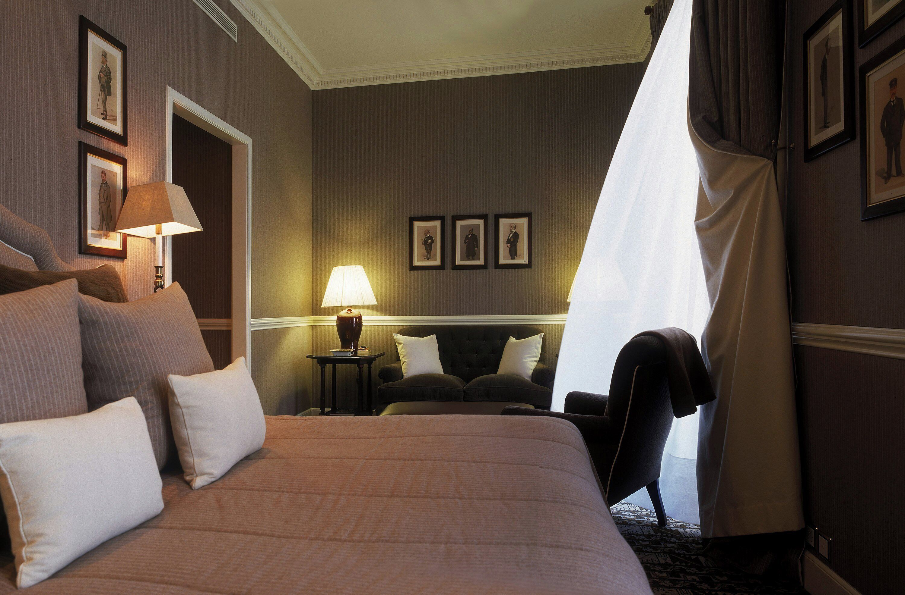 Una habitación de hotel con una cama grande, una zona de estar con una silla, una lámpara y cuadros en la pared.
