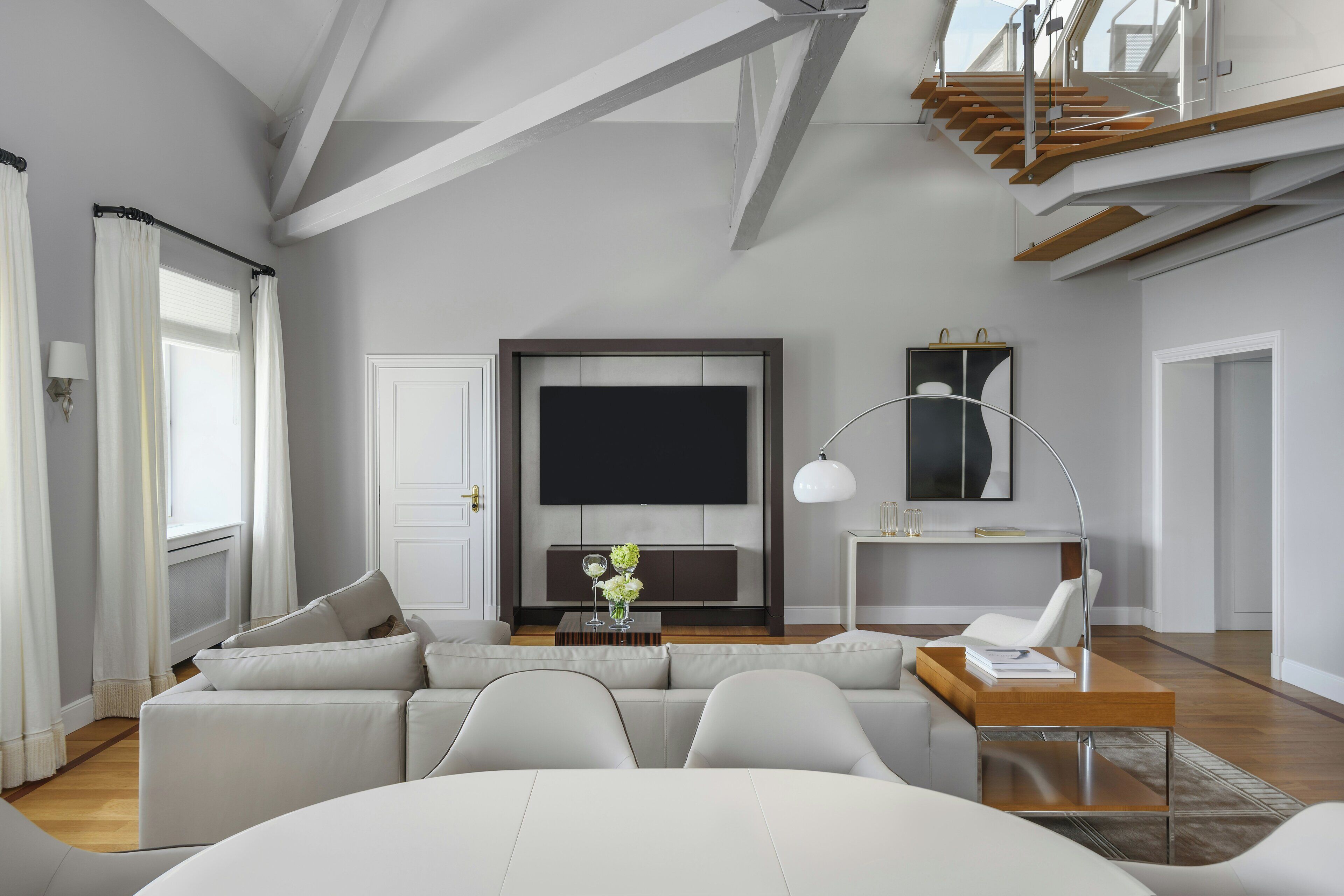 A modern living room with a large flat-screen TV, a sofa set, a coffee table with decorative items, and a staircase leading to an upper level.