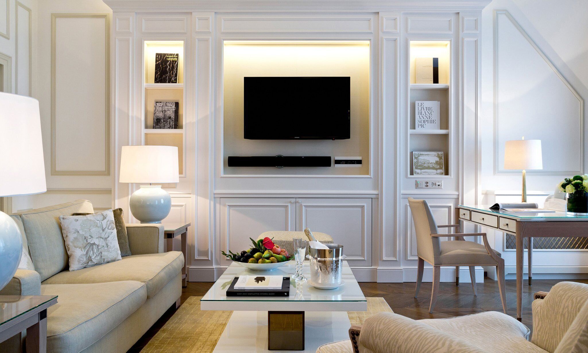 Ein modernes Wohnzimmer mit einem an der Wand befestigten Flachbildfernseher, einem Glastisch, einer Couch, einem Essbereich mit einem Stuhl und einem Bücherregal mit Büchern und Dekorationsobjekten.