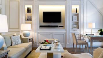 Ein modernes Wohnzimmer mit einem an der Wand befestigten Flachbildfernseher, einem Glastisch, einer Couch, einem Essbereich mit einem Stuhl und einem Bücherregal mit Büchern und Dekorationsobjekten.