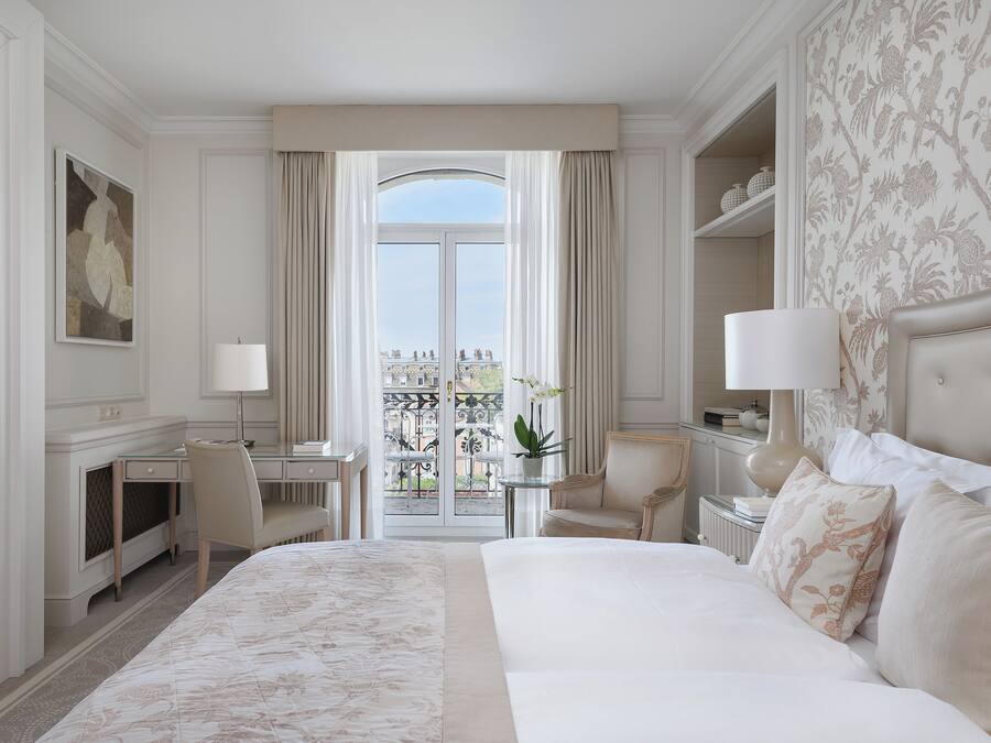 Habitación de hotel con una cama grande, un escritorio, una silla, una mesita de noche con una lámpara y vistas a un balcón con plantas.