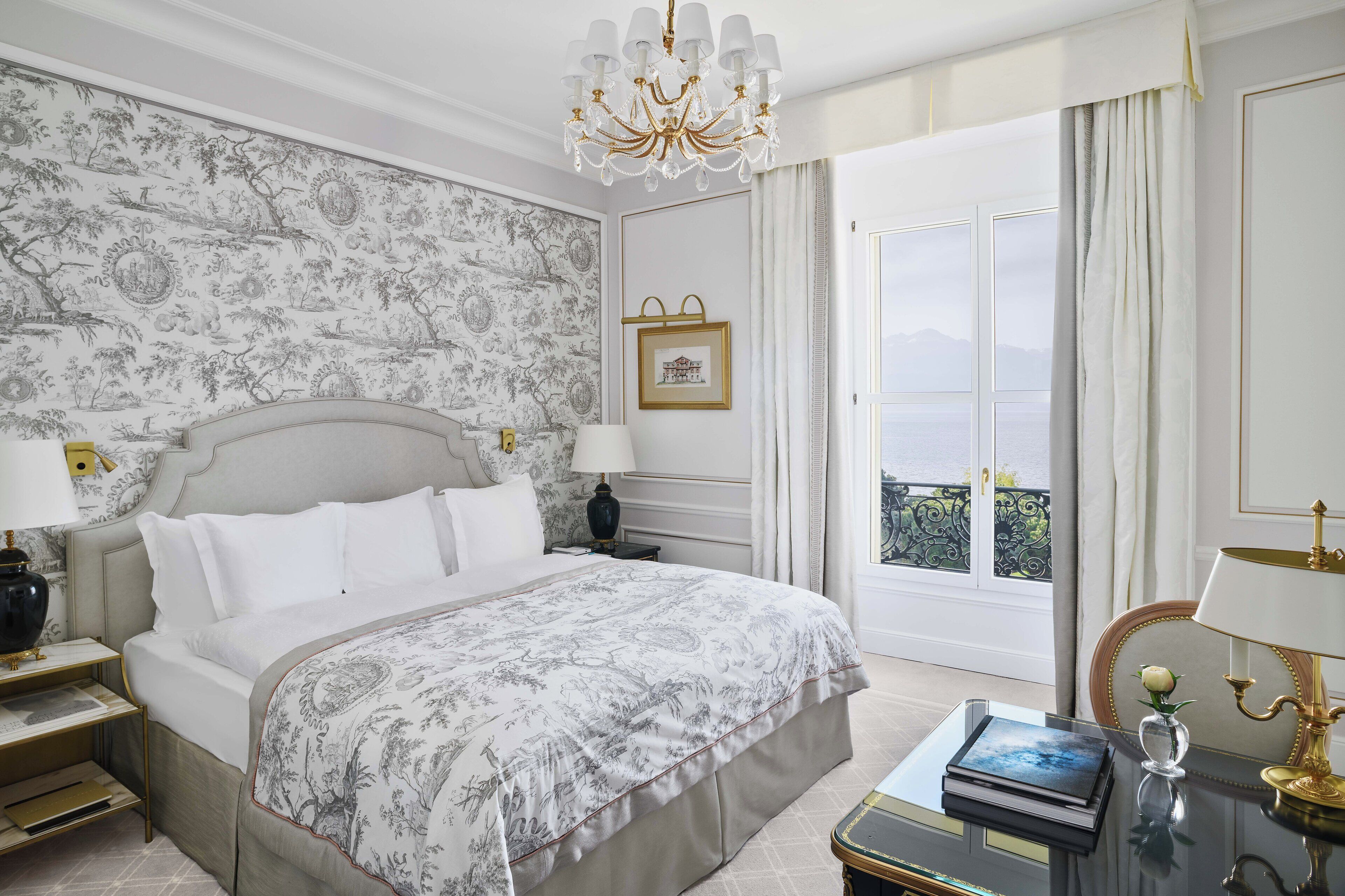 A bedroom with a large bed, a chandelier, a nightstand, a bedside table with a lamp, a framed picture, and a view of the outdoors.