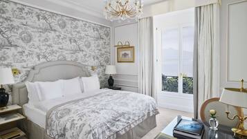 A bedroom with a large bed, a chandelier, a nightstand, a bedside table with a lamp, a framed picture, and a view of the outdoors.
