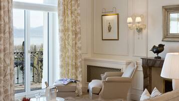 A hotel room with a large bed, a sofa, a chair, a small table, and a chandelier. There is a fireplace and a window with curtains.