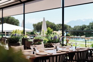 A restaurant with a view of a pool and mountains, featuring a table set with white tableware and potted plants.