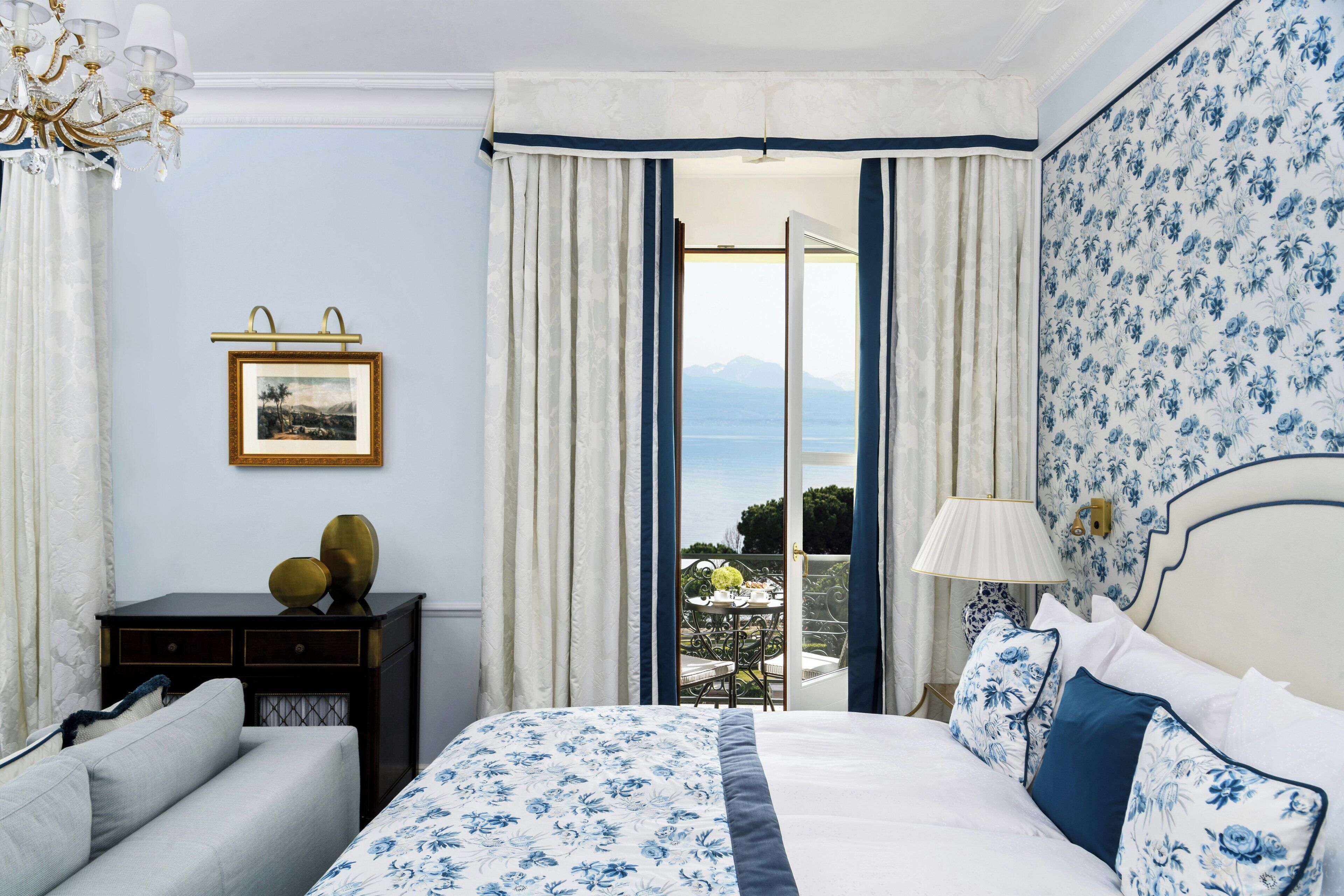 A bedroom with a bed, a desk, a chair, a lamp, and a view of the outdoors.