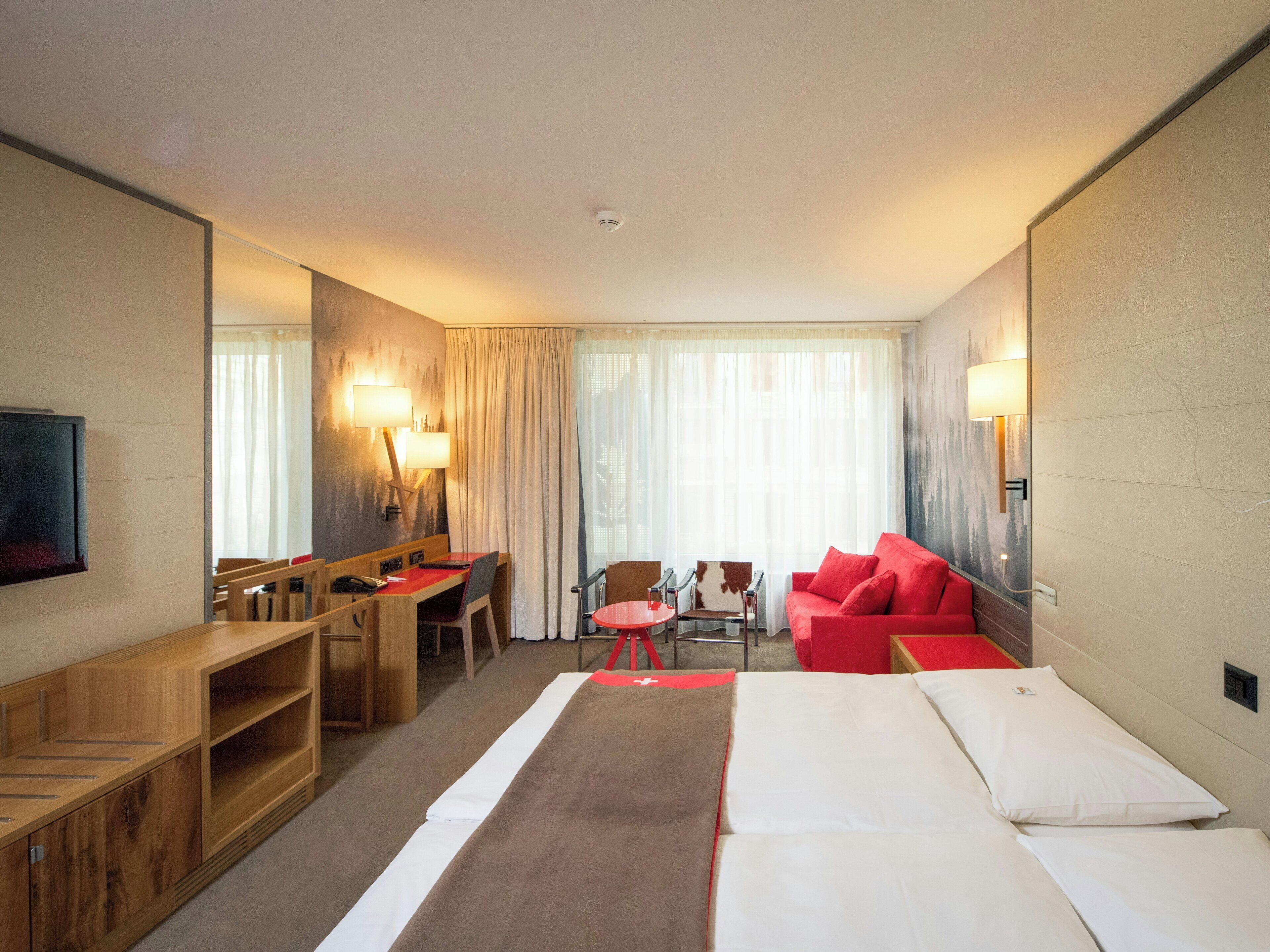 A hotel room with a bed, a desk, a chair, a sofa, a TV, and a window with curtains.