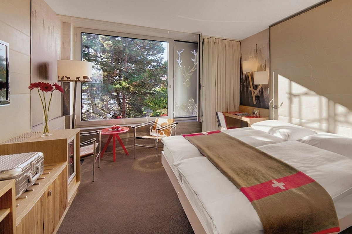 A hotel room with a bed, a desk, a chair, and a window with a view of trees.