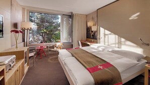 A hotel room with a bed, a desk, a chair, a lamp, and a view of trees through the window.
