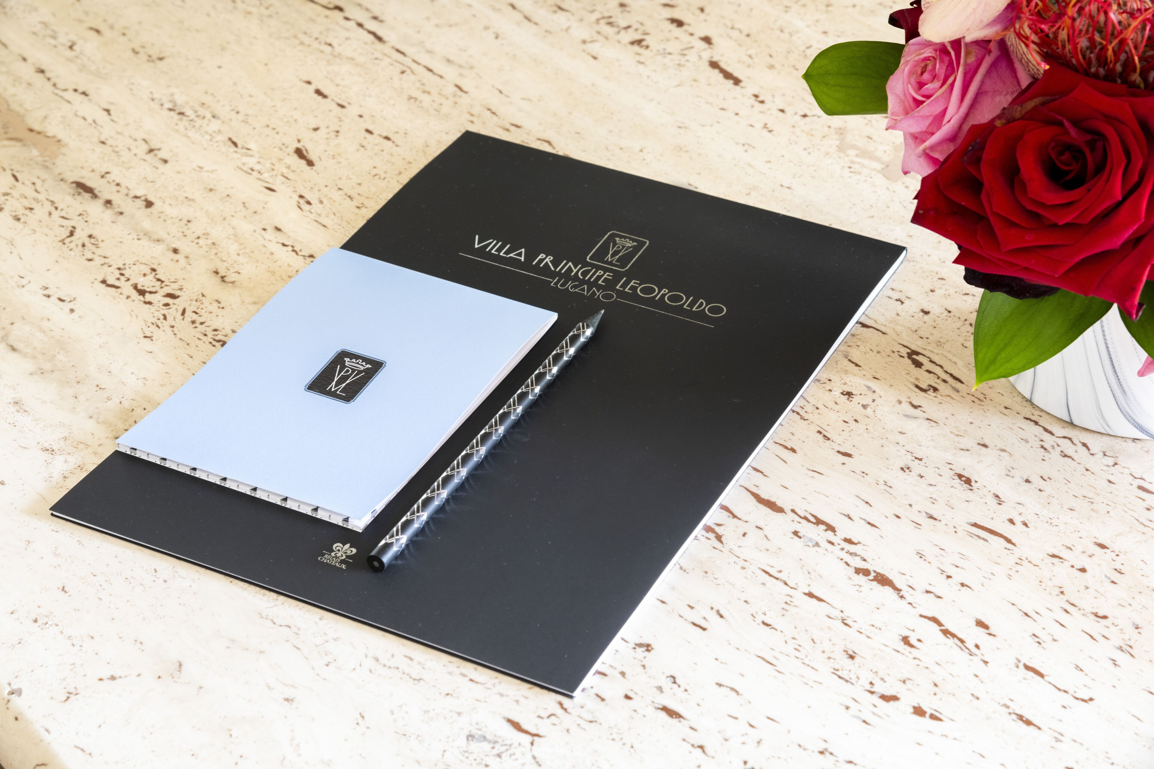 A black folder with a logo and text, a blue notebook, and a pen on a textured surface.