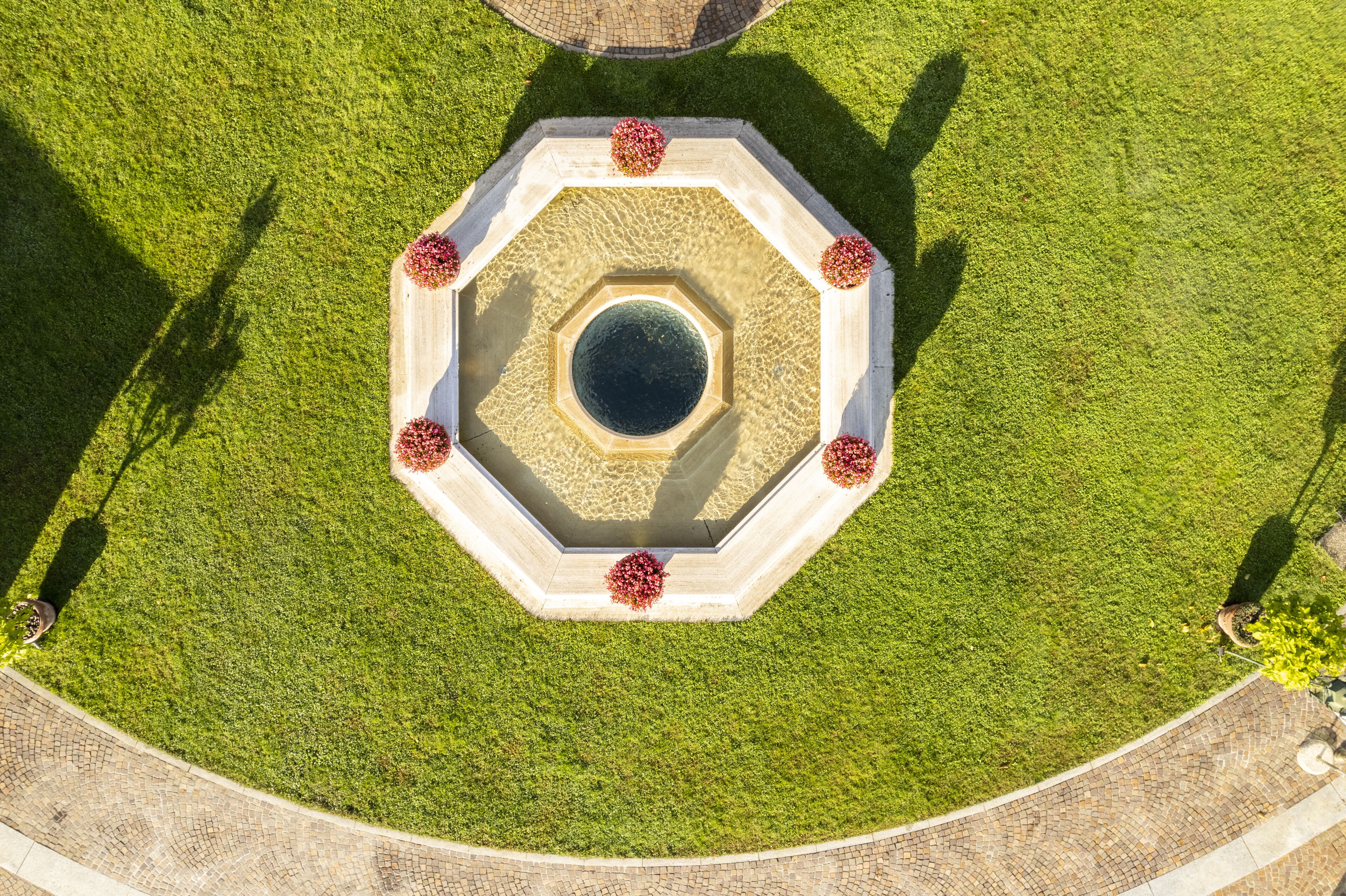 Una fuente octogonal con un elemento acuático central rodeado por un borde de piedra circular, ubicada en un césped cuidado y con un camino pavimentado alrededor.