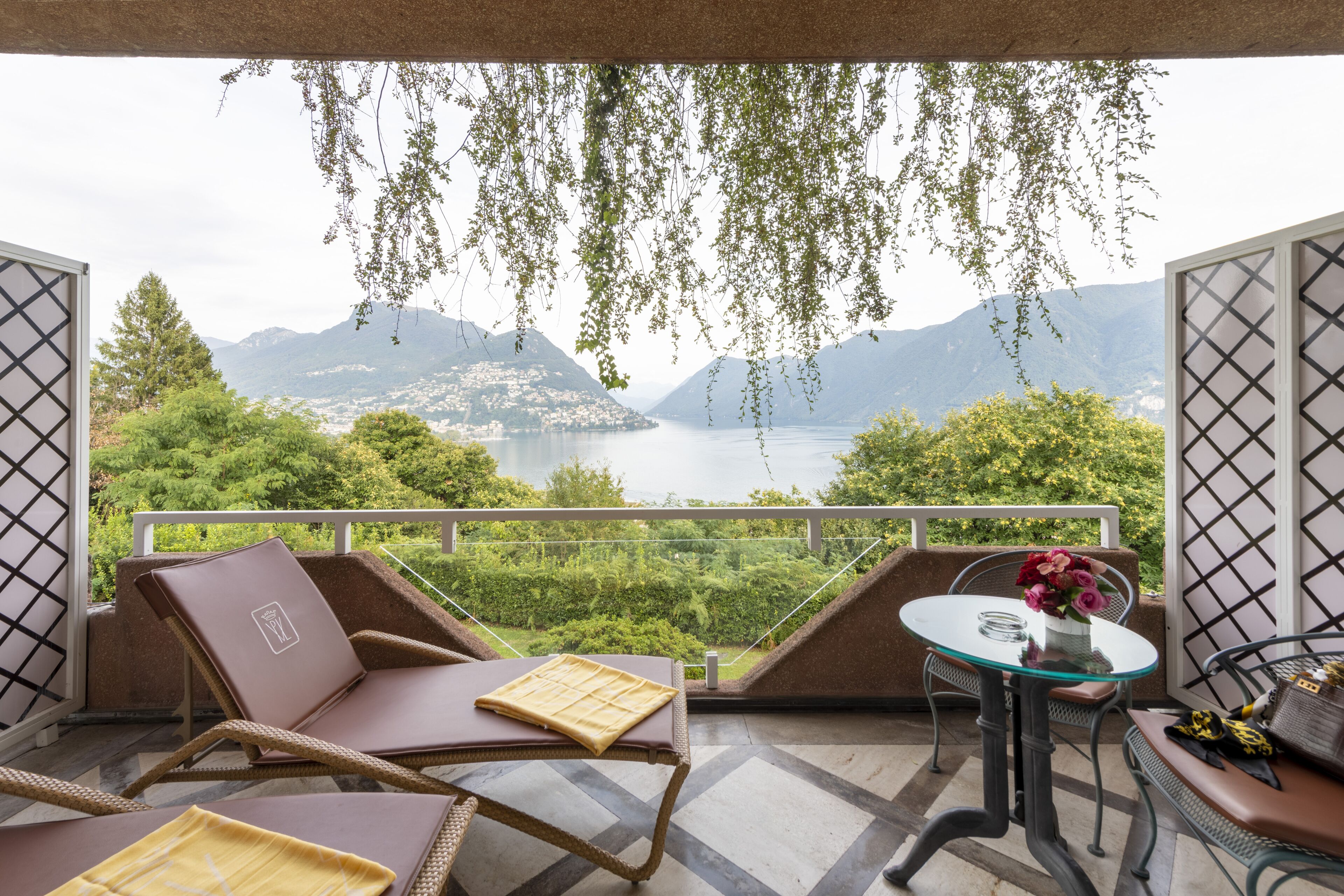 Un balcone con mobili in vimini, un tavolo in vetro e vista sulle montagne e un lago.